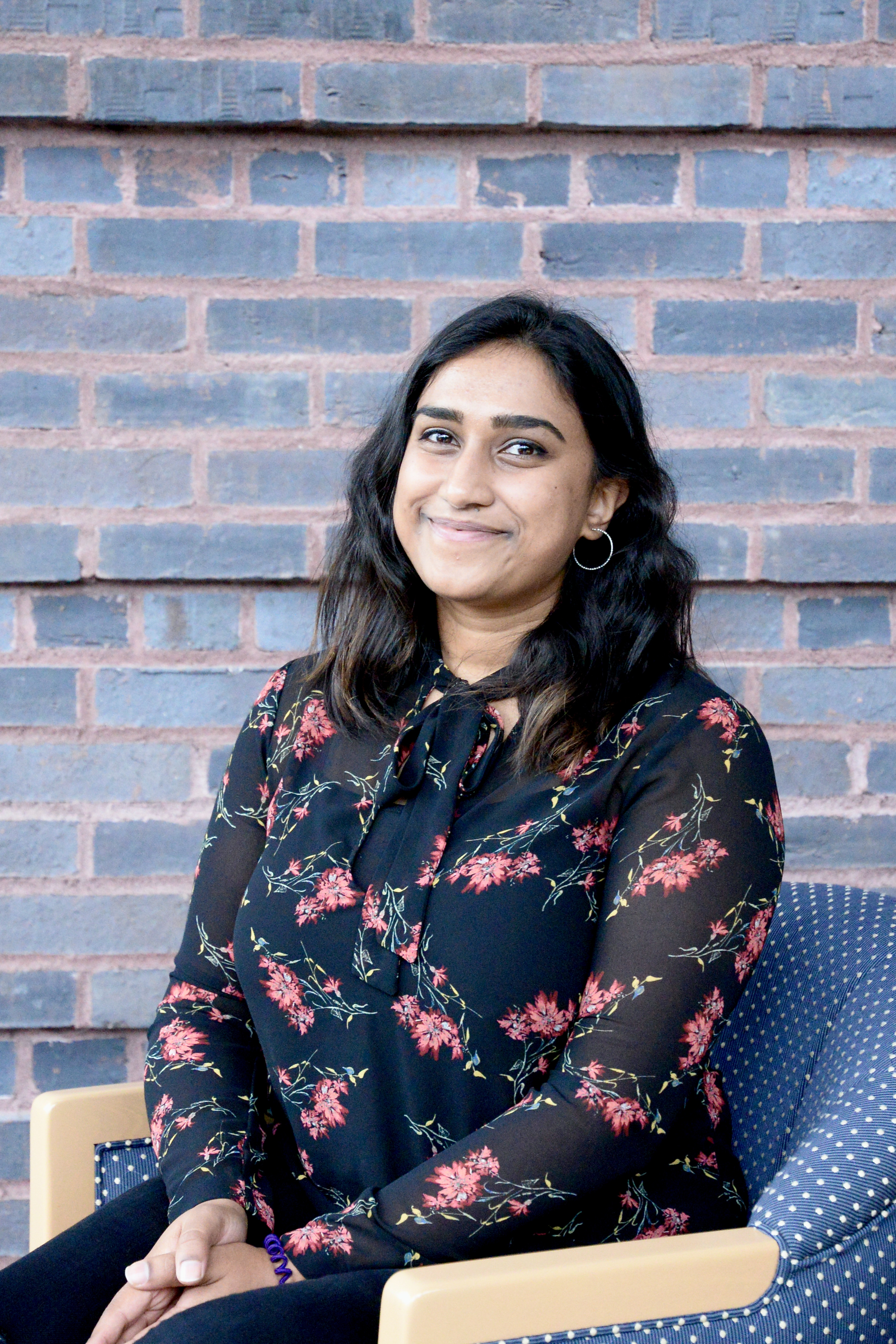 Shivani smiling in a black flower print blouse, seated before a brick wall.